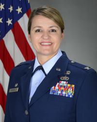 Woman in Air Force Dress Blue Uniform smiling infront of drapped American flag.
